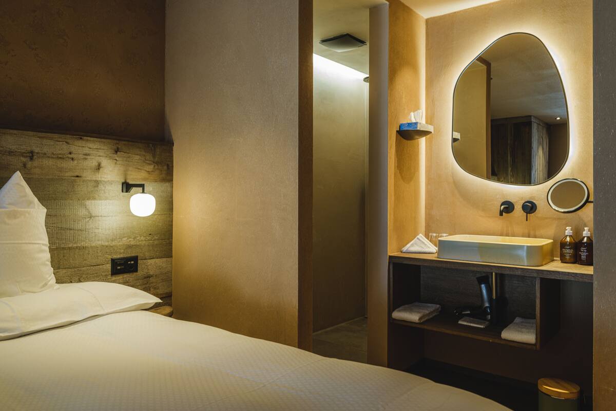 A hotel room with a bed, a wooden headboard, a large mirror, a sink, and a towel rack.