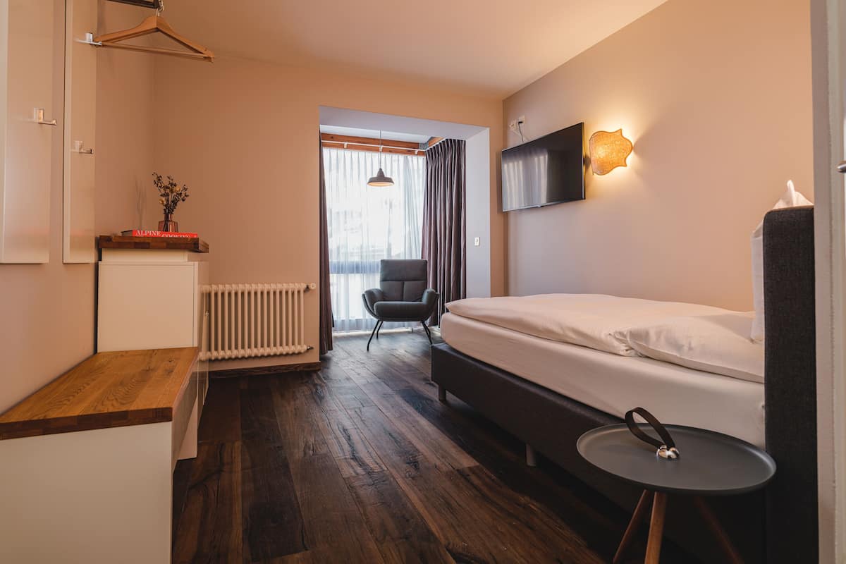 A modern hotel room with a wooden floor, a bed, a small table, a chair, a radiator, and a TV mounted on the wall.