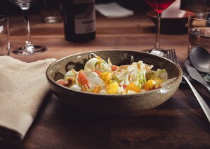 A bowl of salad on a wooden table with a bottle of wine, a glass, and cutlery.