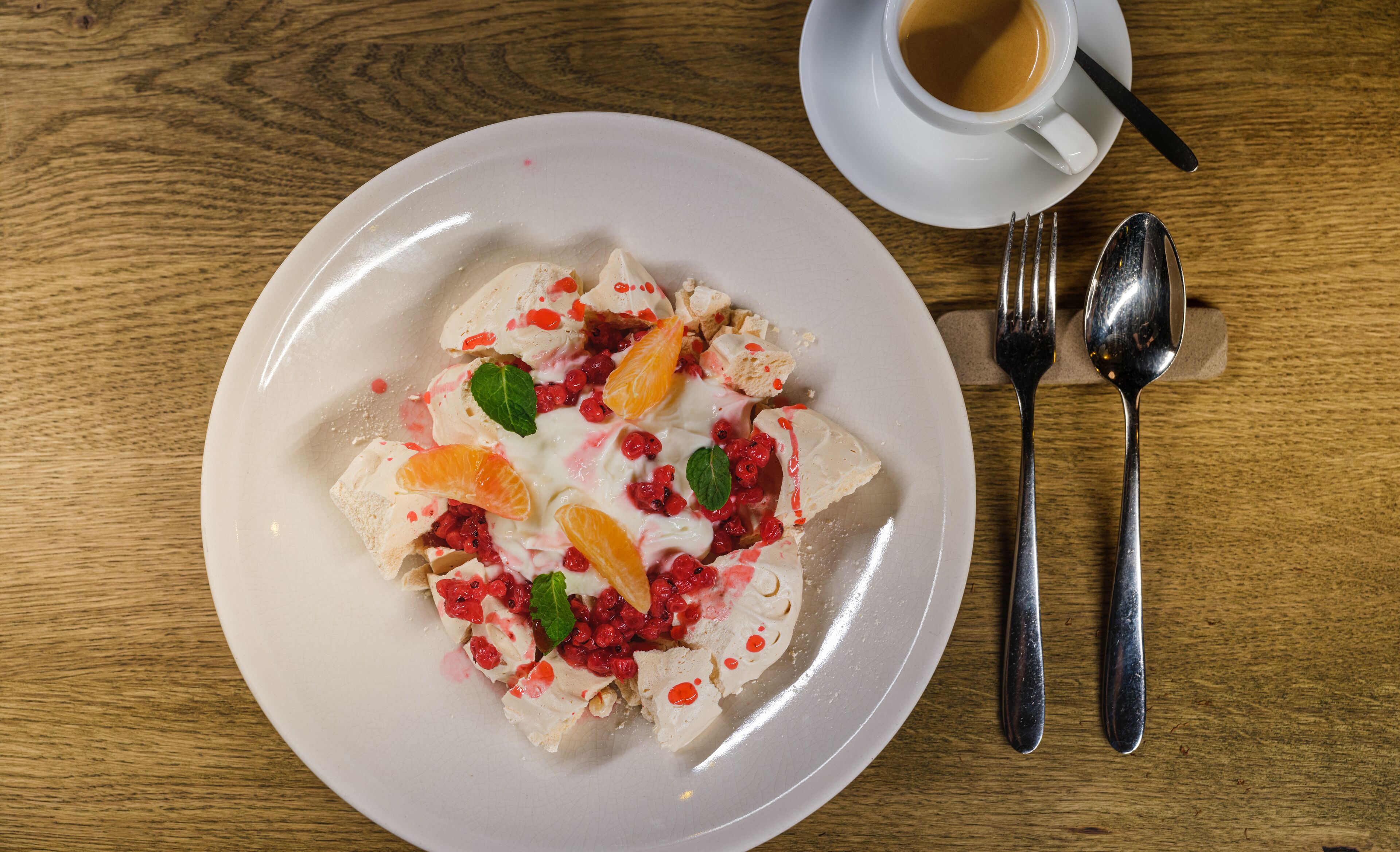 Un plato de postre con fruta y crema sobre una mesa de madera, acompañado de una taza de café y cubiertos.