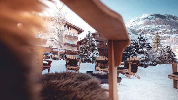 Un espace extérieur enneigé avec des chaises et des tables en bois, un arbre recouvert de neige et un bâtiment doté de balcons.