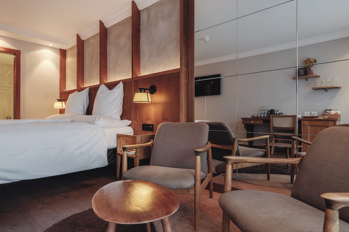 A hotel room with a large bed, two armchairs, a small wooden table, a TV, and a minibar.