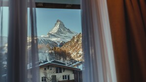 Une vue sur une montagne, aperçue à travers une fenêtre ornée d’un rideau, avec un sommet enneigé et des arbres environnants.