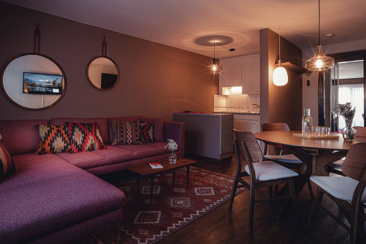 A modern living room with a sofa, coffee table, and dining area. There are two round mirrors on the wall, a TV mounted above the sofa, and a kitchen area in the background.
