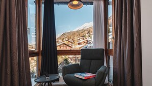 Une pièce chaleureuse donnant sur un village de montagne enneigé, avec un fauteuil confortable, une petite table et un livre intitulé « Cuisine alpine ».