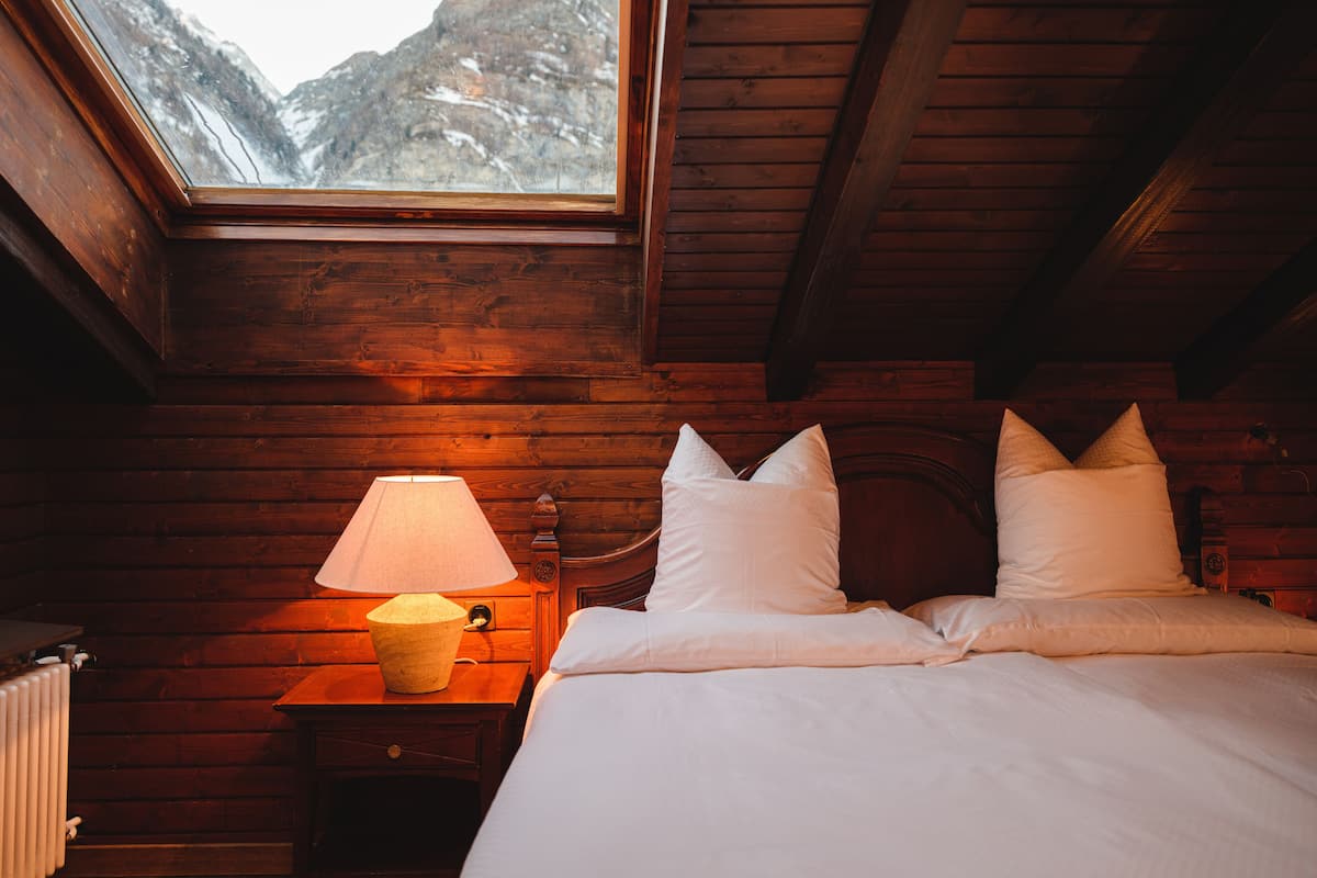 A wooden cabin room with a bed, a nightstand with a lamp, and a window overlooking a mountainous landscape.