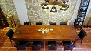 Une table à manger en bois, entourée de six chaises, adossée à un mur en pierre et complétée par une bibliothèque contenant divers objets.