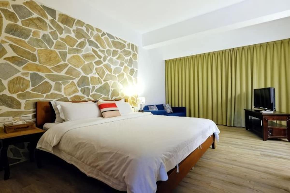 Habitación de hotel con pared de piedra, cama con ropa blanca, suelo de madera, televisor sobre un soporte y cortina verde.