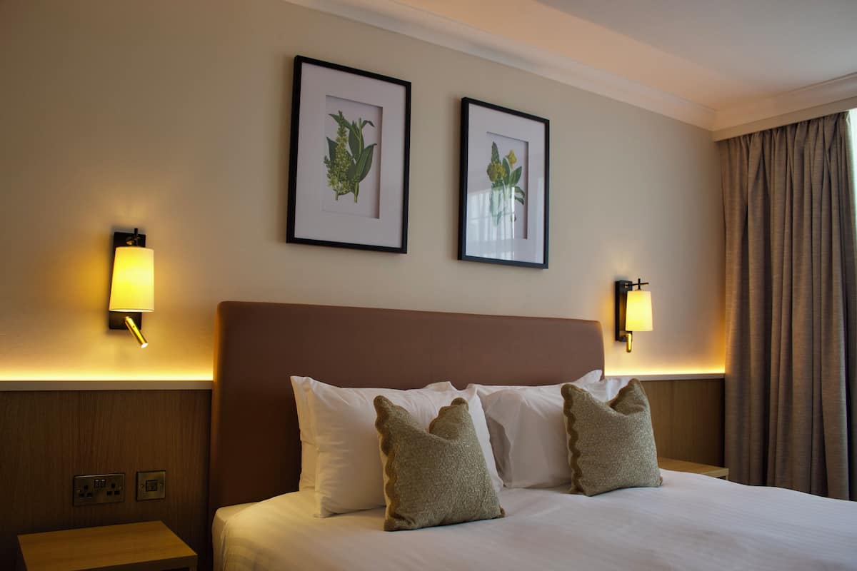 Habitación de hotel con una cama grande, dos lámparas fijadas a la pared y dos impresiones botánicas enmarcadas.
