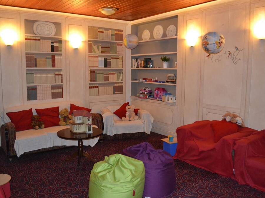 Um quarto acolhedor com sofá, cadeiras e puffs. Há estantes com livros e objetos decorativos.