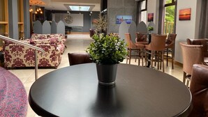 Un moderno vestíbulo de hotel con un techo alto, una zona de estar central con sofás y sillones, una mesa redonda con una planta en maceta y un piano al fondo.
