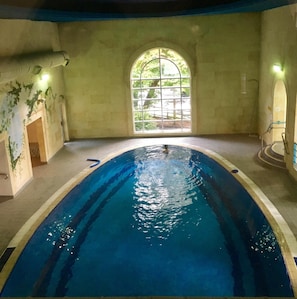 An indoor swimming pool with clear water, a tiled floor, and a large arched window allowing natural light.
