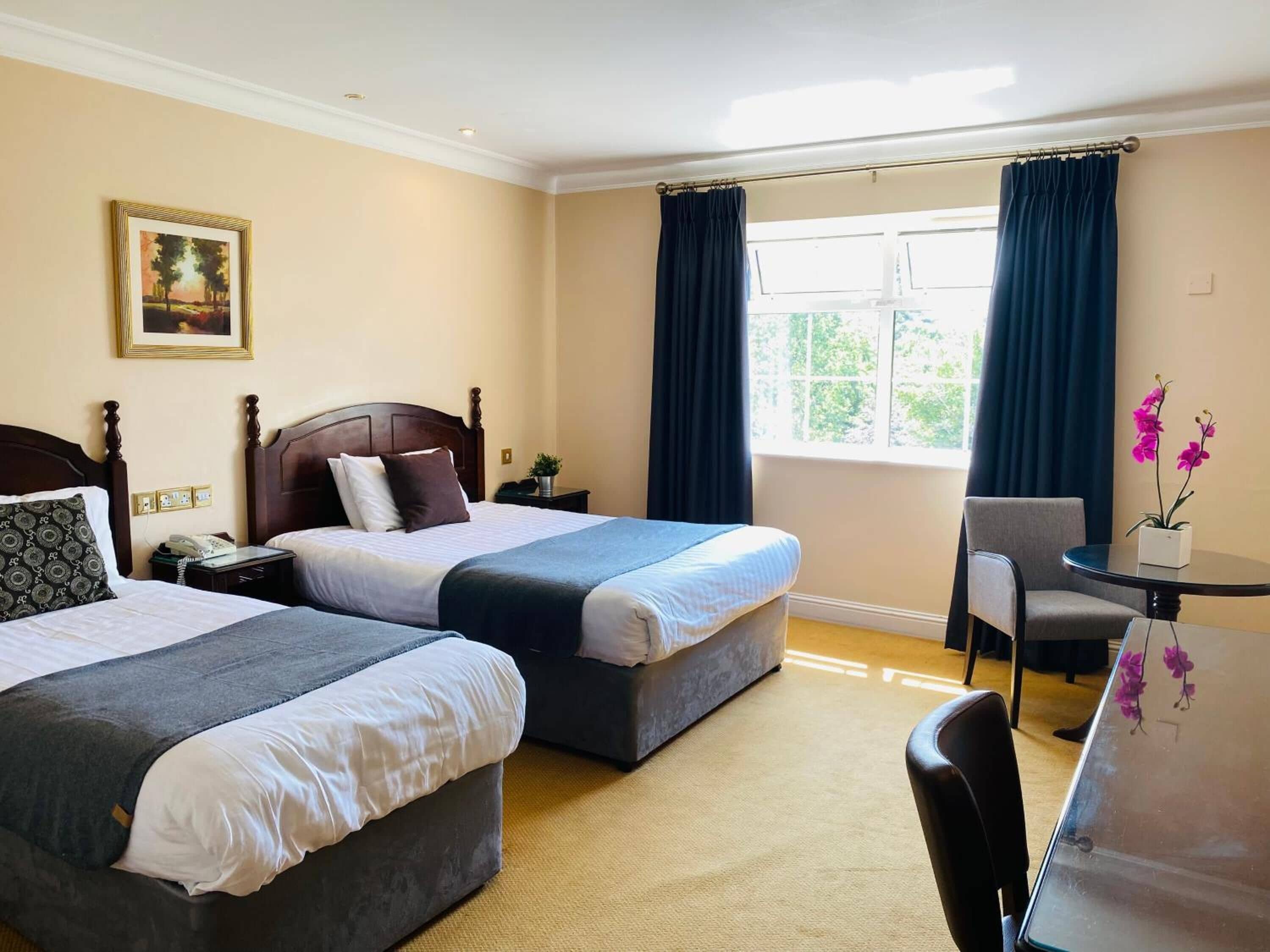 A hotel room with two beds, a desk, a chair, a window with curtains, and a painting on the wall.