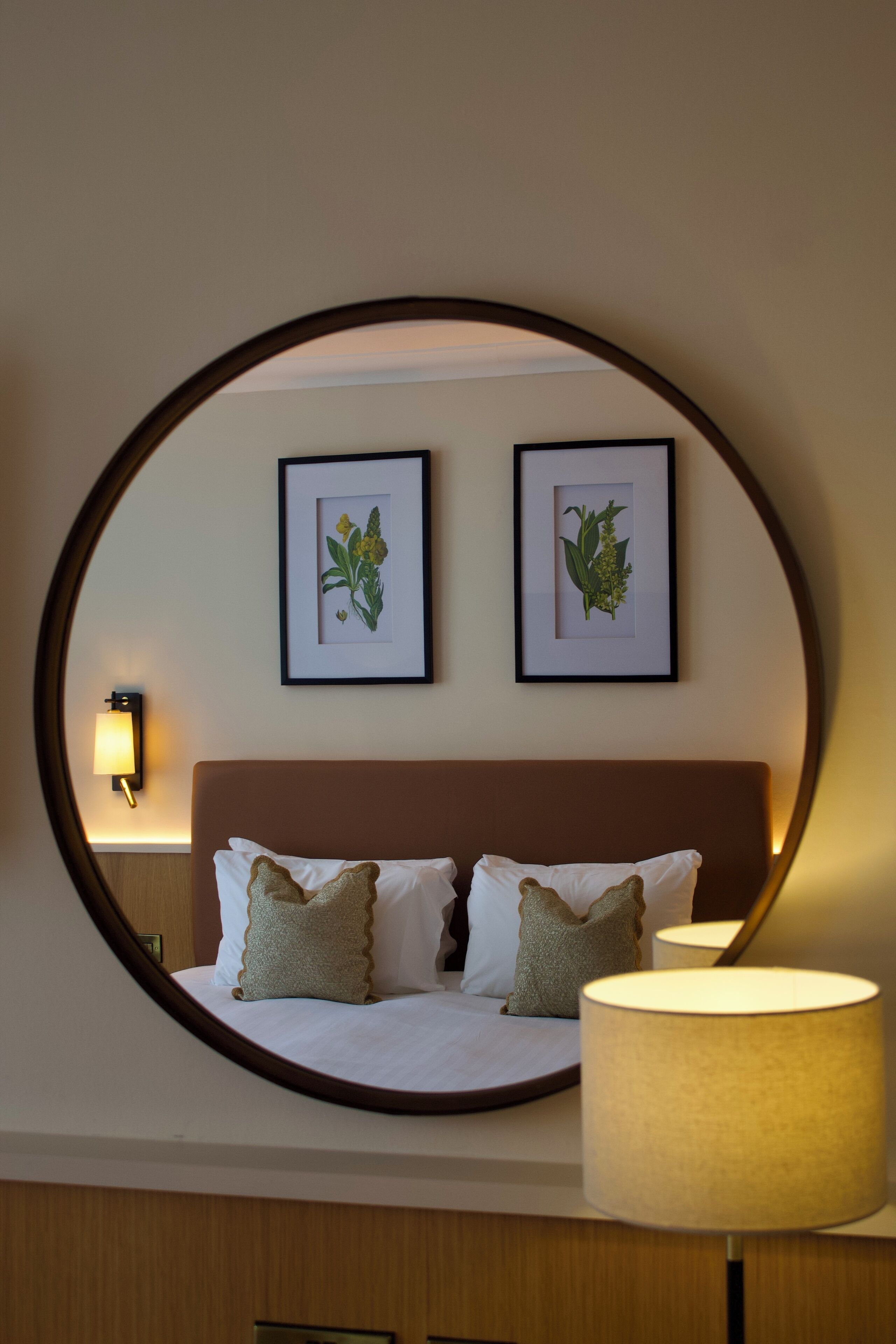A bedroom with a bed, pillows, a bedside lamp, and two framed botanical prints on the wall.