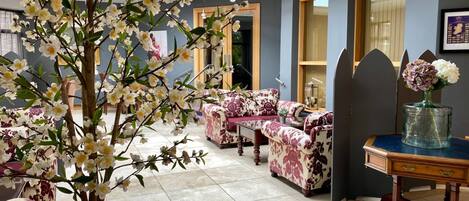 Una habitación con un árbol artificial grande, sofás con estampado floral y un florero de vidrio sobre una mesa de madera.