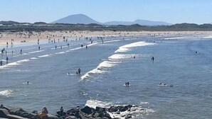 Una playa con gente nadando y caminando por la orilla, con montañas al fondo.