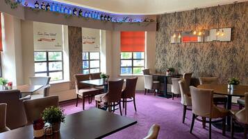 En restaurant i et hotel med flere borde og stole, store vinduer og et dekorativt loft.