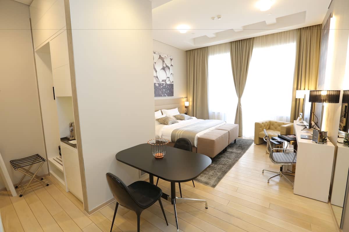 A modern hotel room with a neatly arranged bed, a dining area with a table and chairs, a desk with a chair, and a large window with curtains.