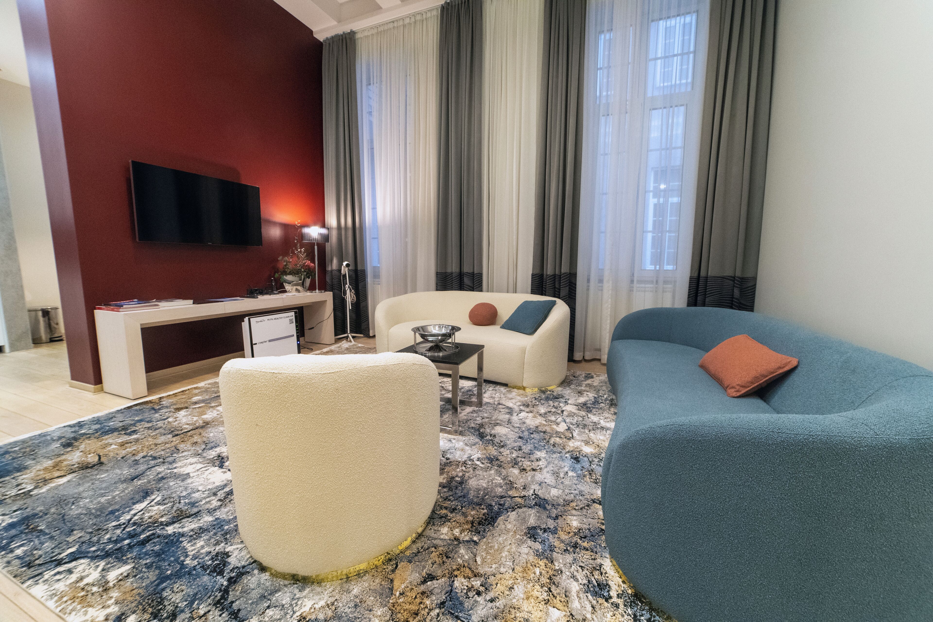 A modern living room with a sofa, armchair, and ottoman, a flat-screen TV mounted on a red wall, and a dining area with a table and chairs.