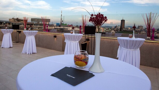 A rooftop terrace with round tables set for dining, a white vase with flowers, and a cityscape view in the background.