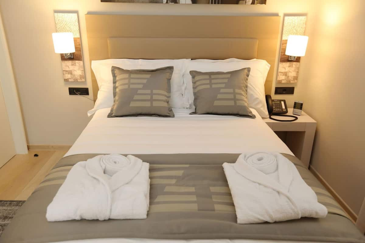 A hotel room with a bed, two pillows, a bedside phone, and two rolled towels on the bed.