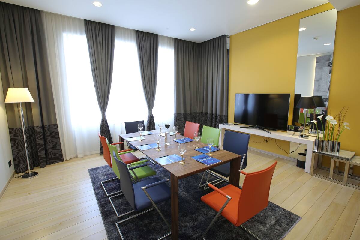 A modern hotel room with a wooden dining table, colorful chairs, a flat-screen TV, and a large mirror.