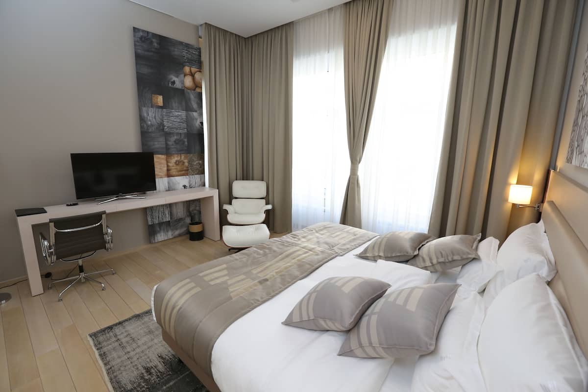A hotel room with a bed, a desk with a TV, a chair, and a large window with curtains.
