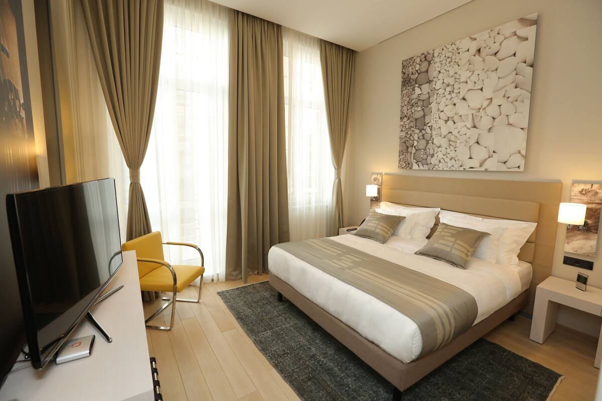 A hotel room with a bed, a TV, a chair, and a painting on the wall.