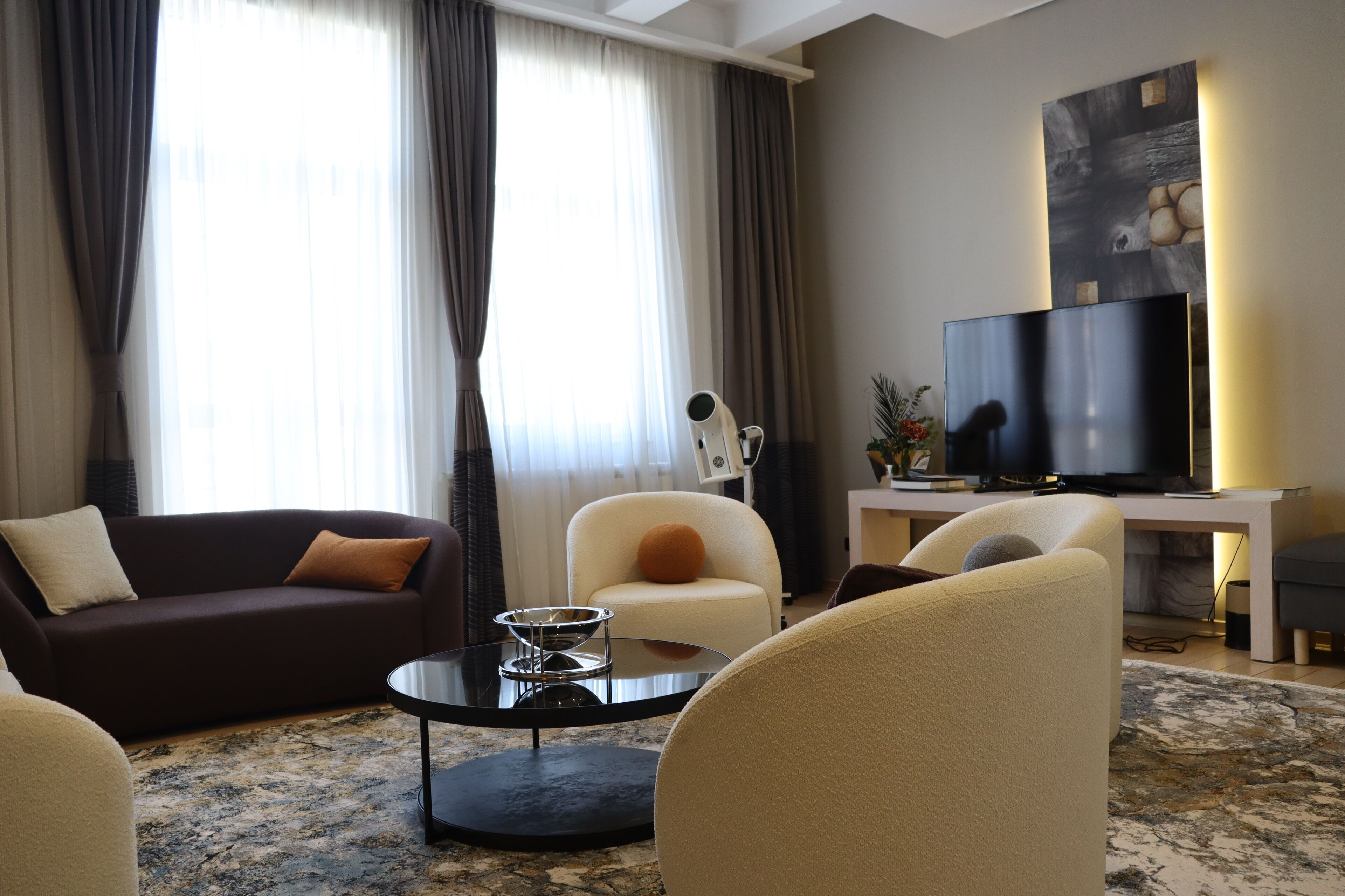 A modern living room with a dark brown sofa, a cream-colored armchair, a round coffee table, and a flat-screen TV.