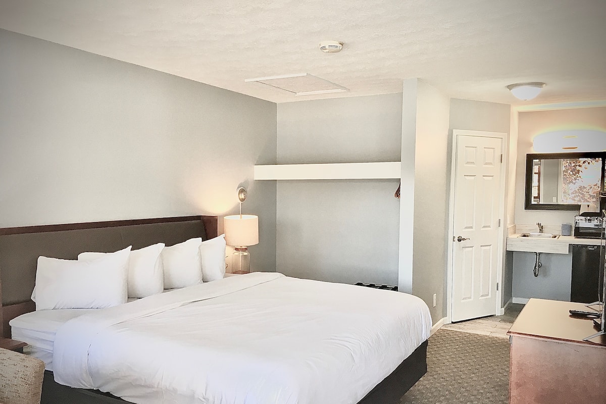 A hotel room with a large bed, a bedside lamp, a built-in shelf, a bathroom with a sink and mirror, and a door leading to another room.