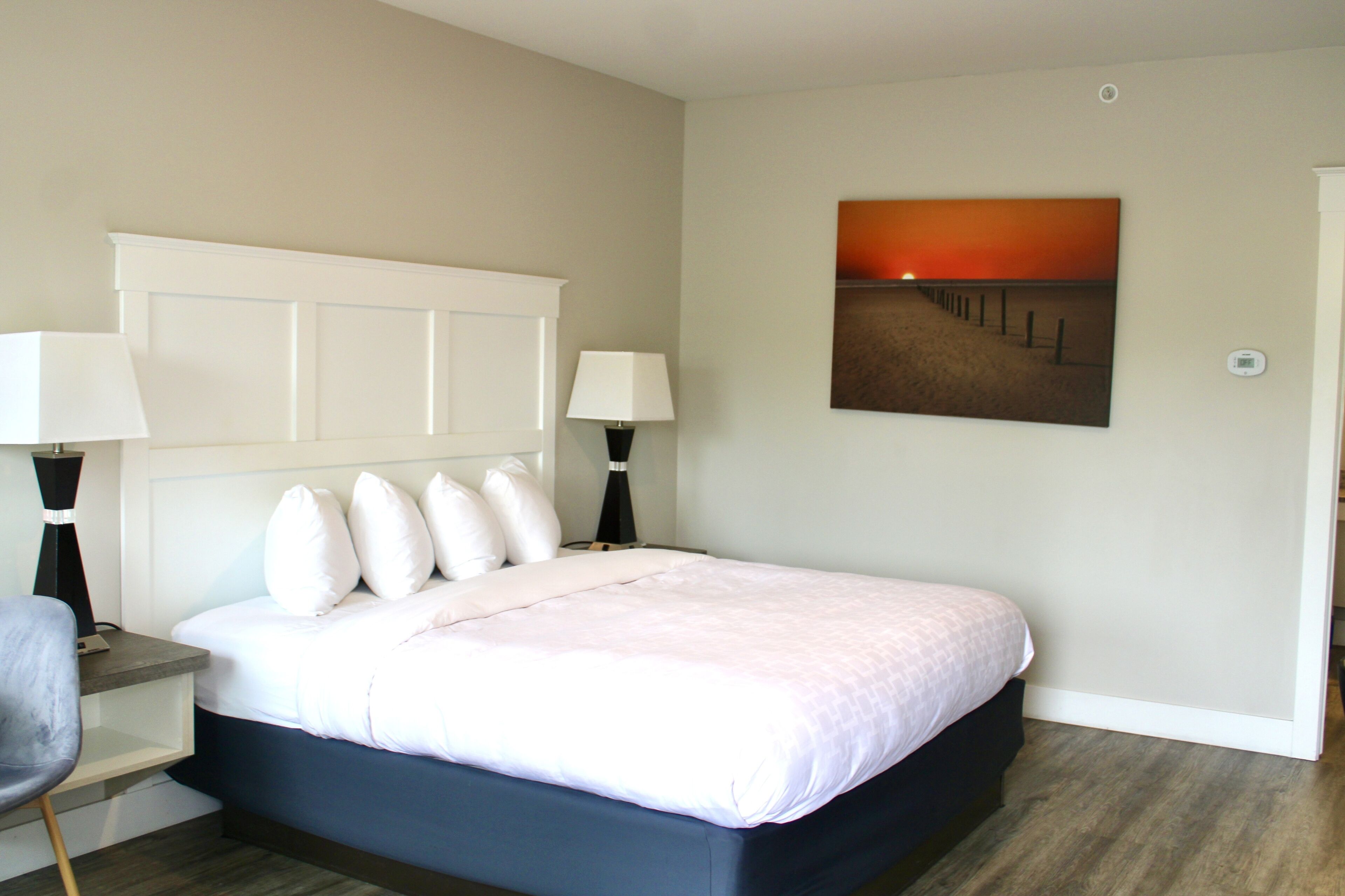 A neatly arranged bedroom with a bed, two bedside tables with lamps, a chair, and a painting on the wall.
