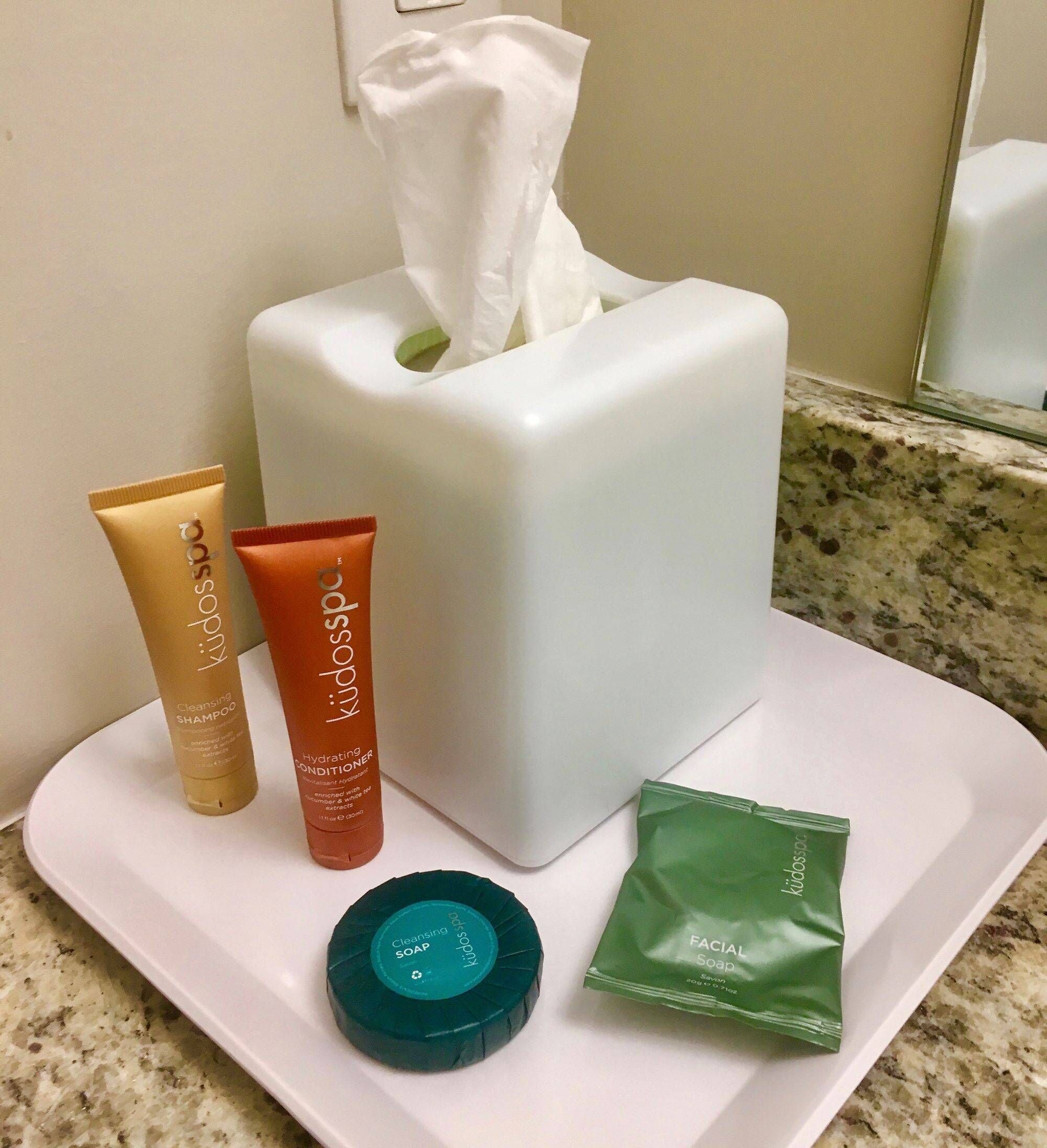 A bathroom counter with a tissue box, two bottles of kids' soap, a facial soap packet, and a round soap bar.