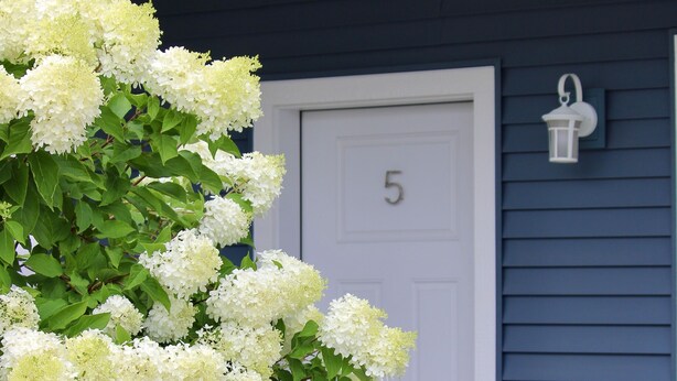 Una casa con puerta blanca numerada con el número 5, fachada azul oscuro y un porche blanco con una silla de mimbre.