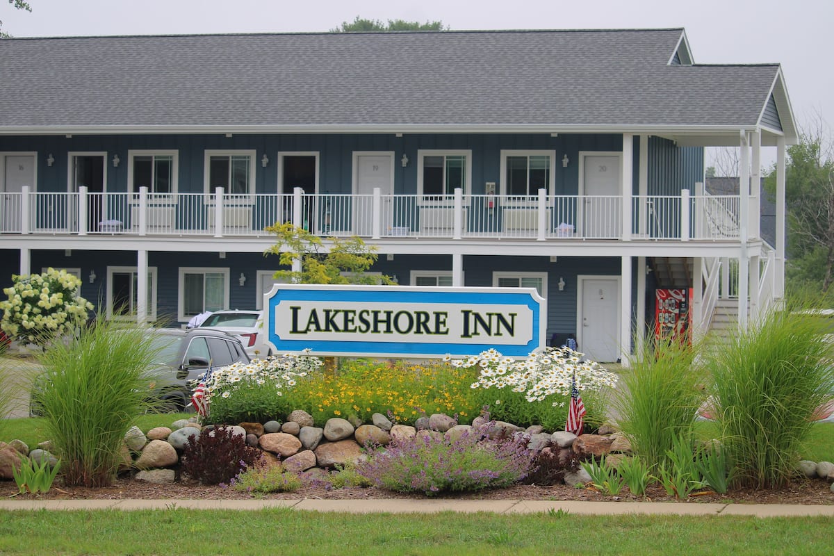 A two-story Lakeshore Inn with a balcony on the upper floor, a landscaped front yard with rocks and plants, and an American flag.