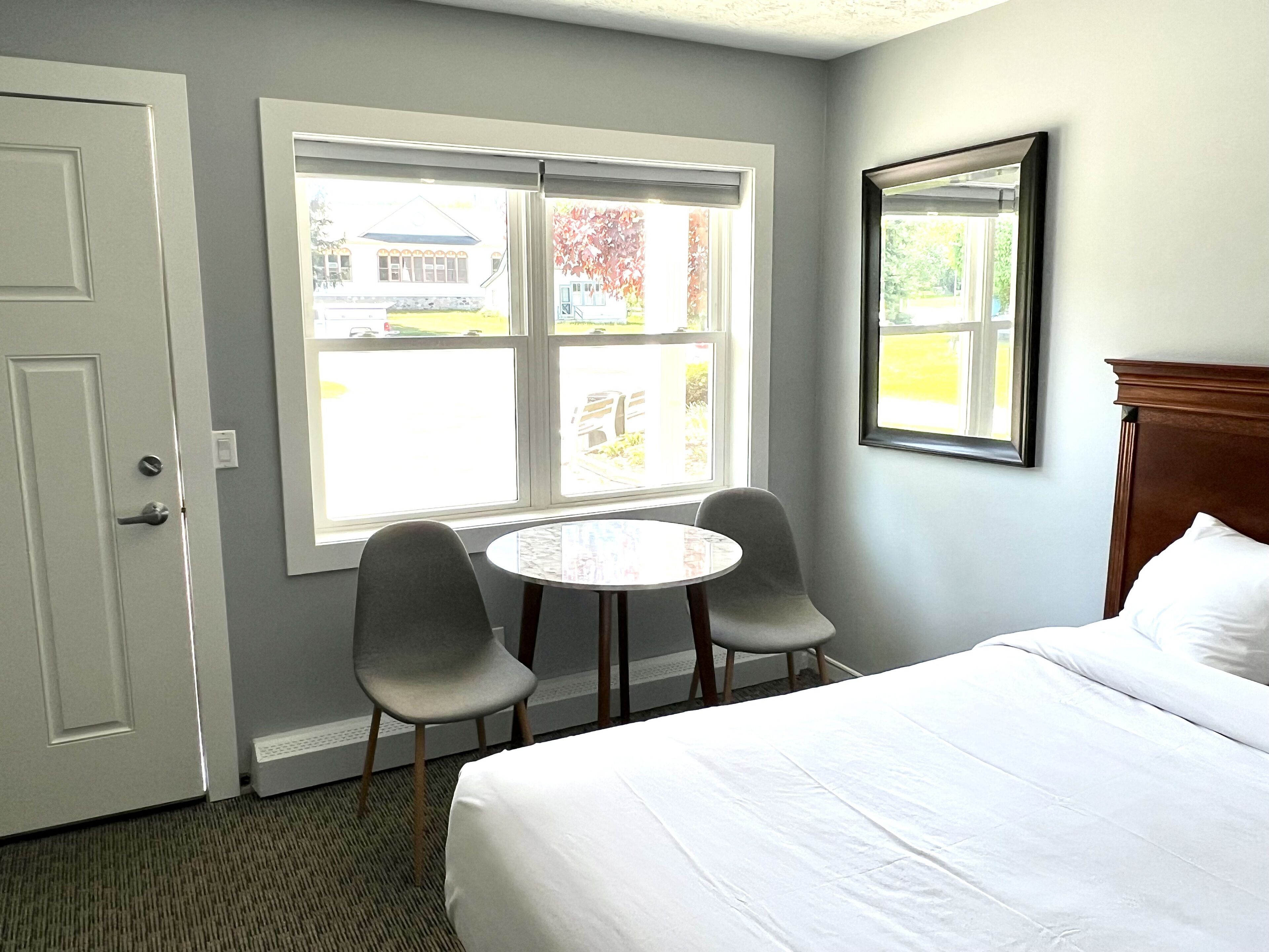 A hotel room with a bed, two chairs, a small table, and a window with a view of a building and trees.