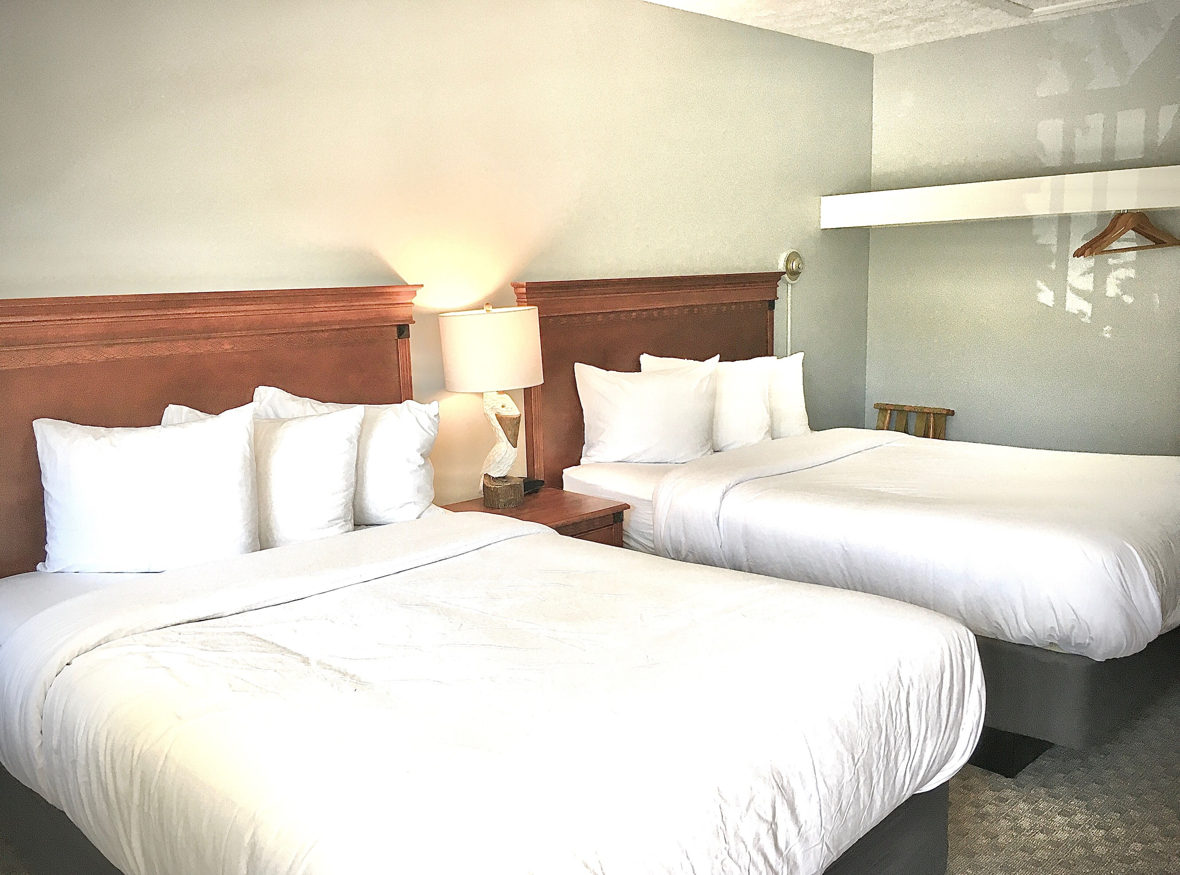 A hotel room with two beds, a wooden headboard, a nightstand with a lamp, and a wall-mounted coat rack.