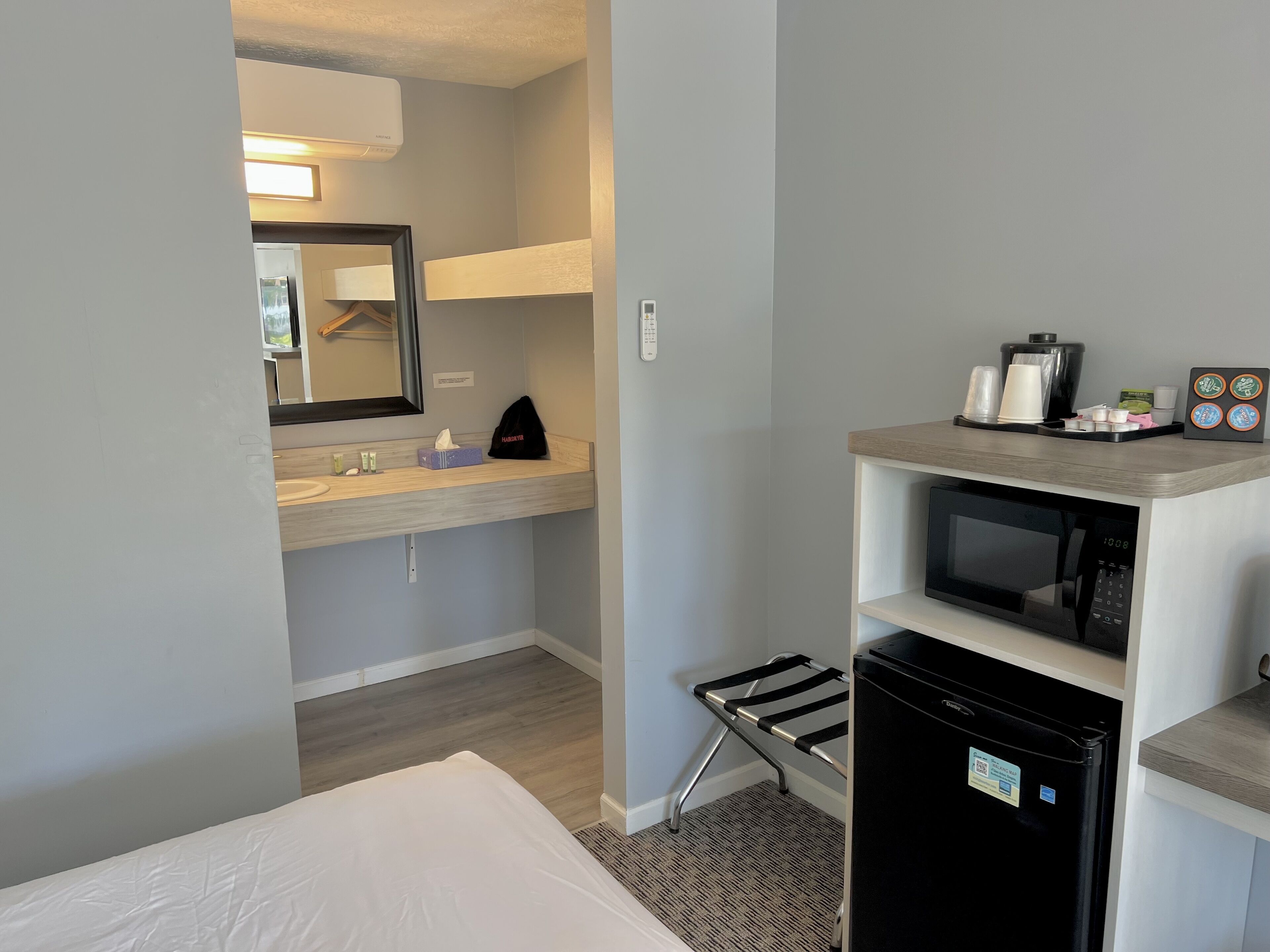 Habitació d'hotel amb un llit, un bany amb un lavabo i mirall, un microones, un frigorífic i una petita estant amb productes de tocador.