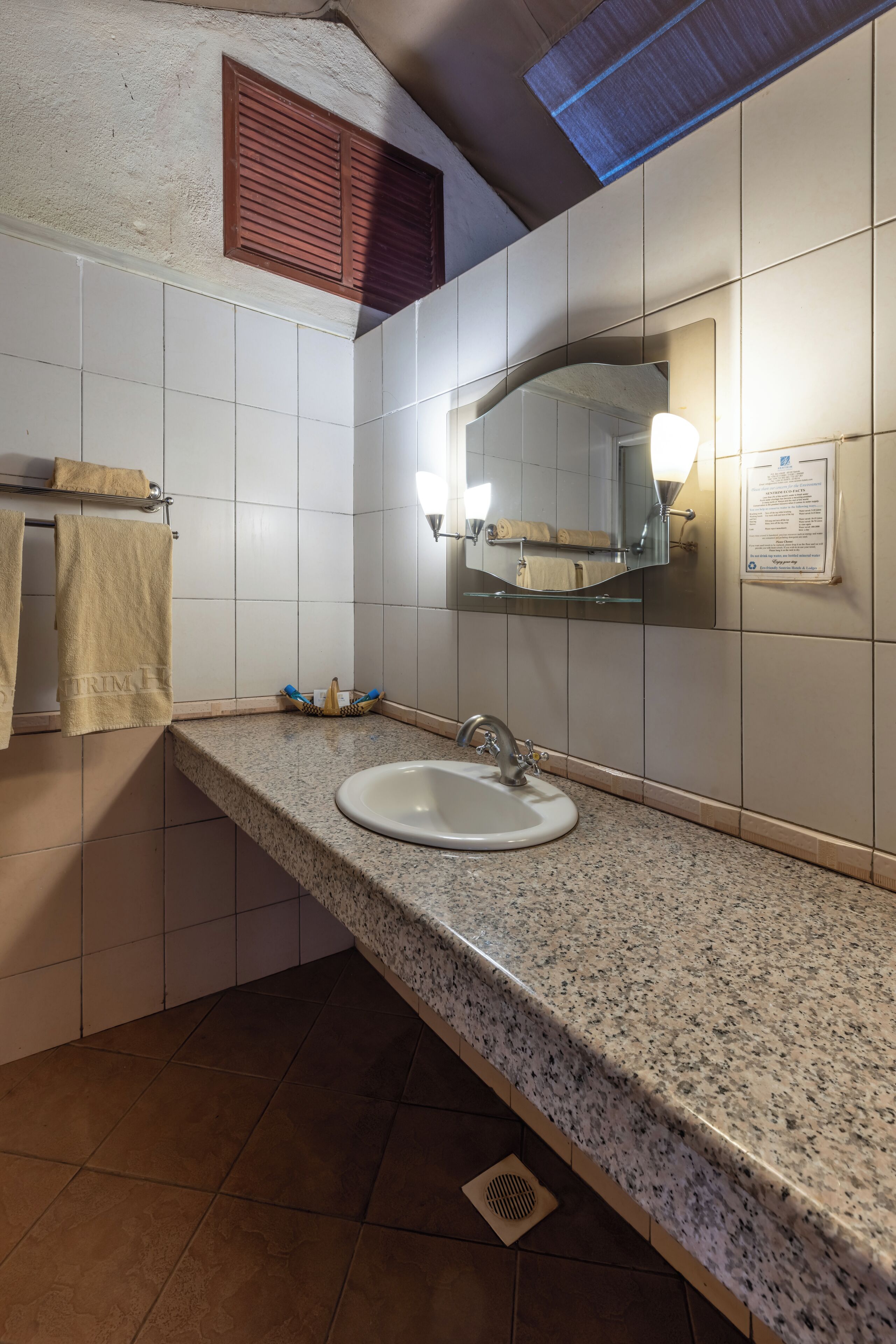 Une salle de bain avec un comptoir en granit, un lavabo, un miroir avec éclairage intégré, un porte-serviettes et une bouche d’aération.