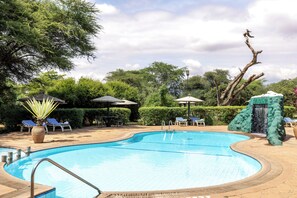 A swimming pool with lounge chairs and umbrellas, surrounded by greenery and a waterfall feature.