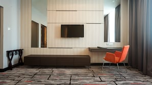 Een moderne hotelkamer met een grote flatscreen televisie aan de muur, een zithoek met een bank en een enkele oranje fauteuil.