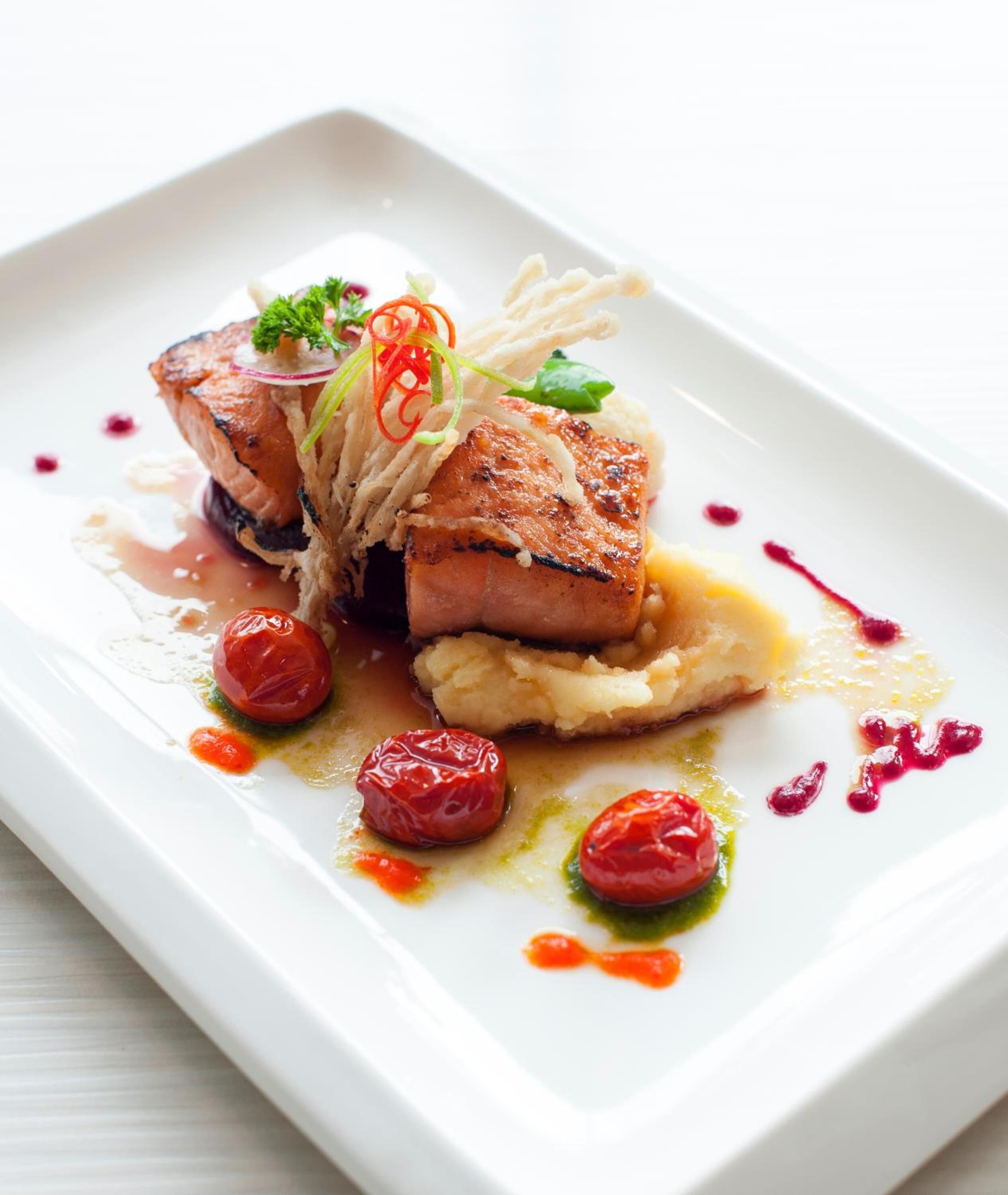 A white plate with a dish consisting of a piece of meat, possibly pork, topped with a garnish and surrounded by small red berries and a sauce.