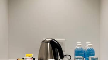 A hotel room with a white wall, a coffee maker, two cups, a kettle, and three water bottles.