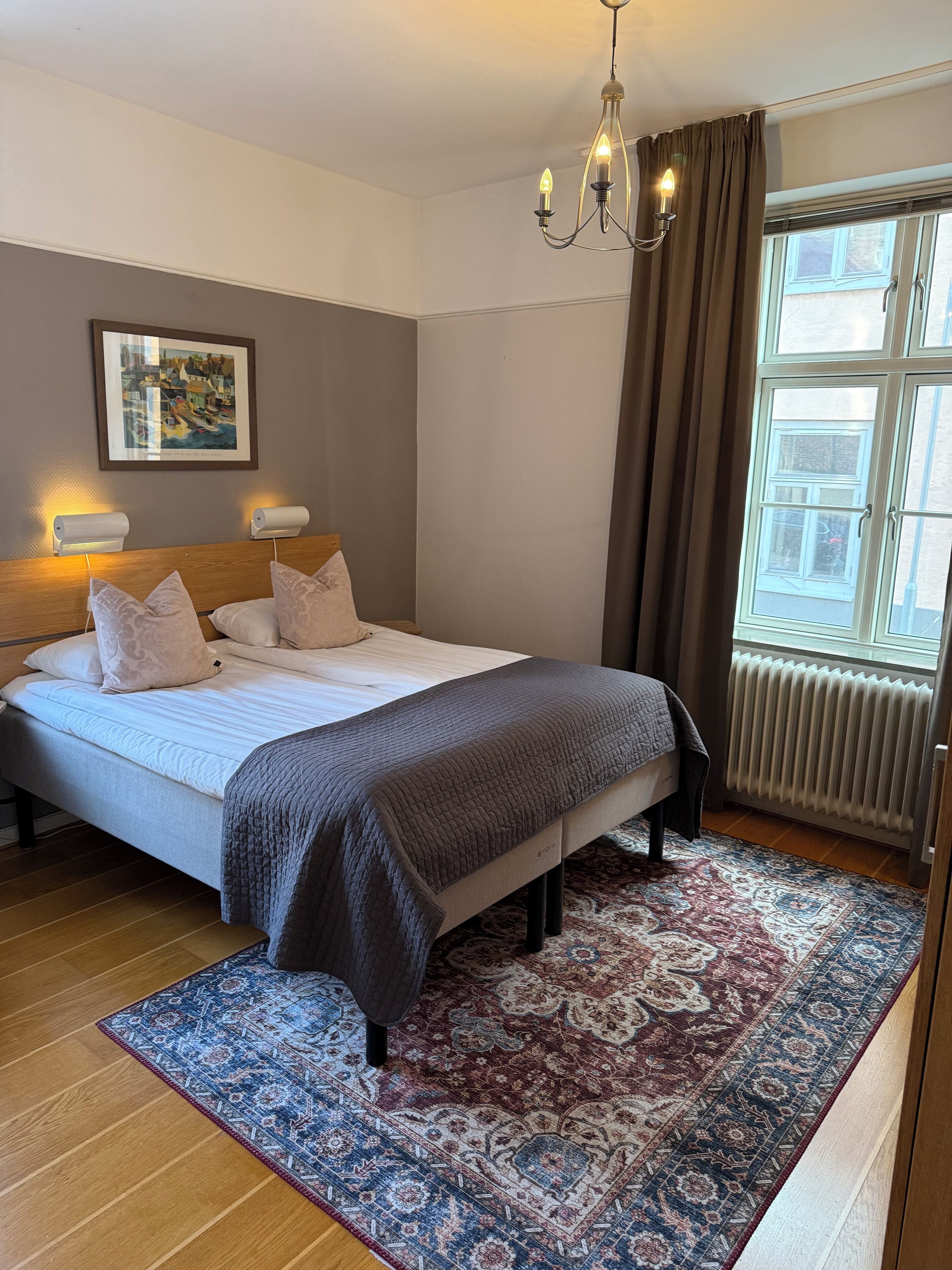 Ett välordnat sovrum med en säng, sänglampor, en mönstrad matta och ett fönster med gardiner.
