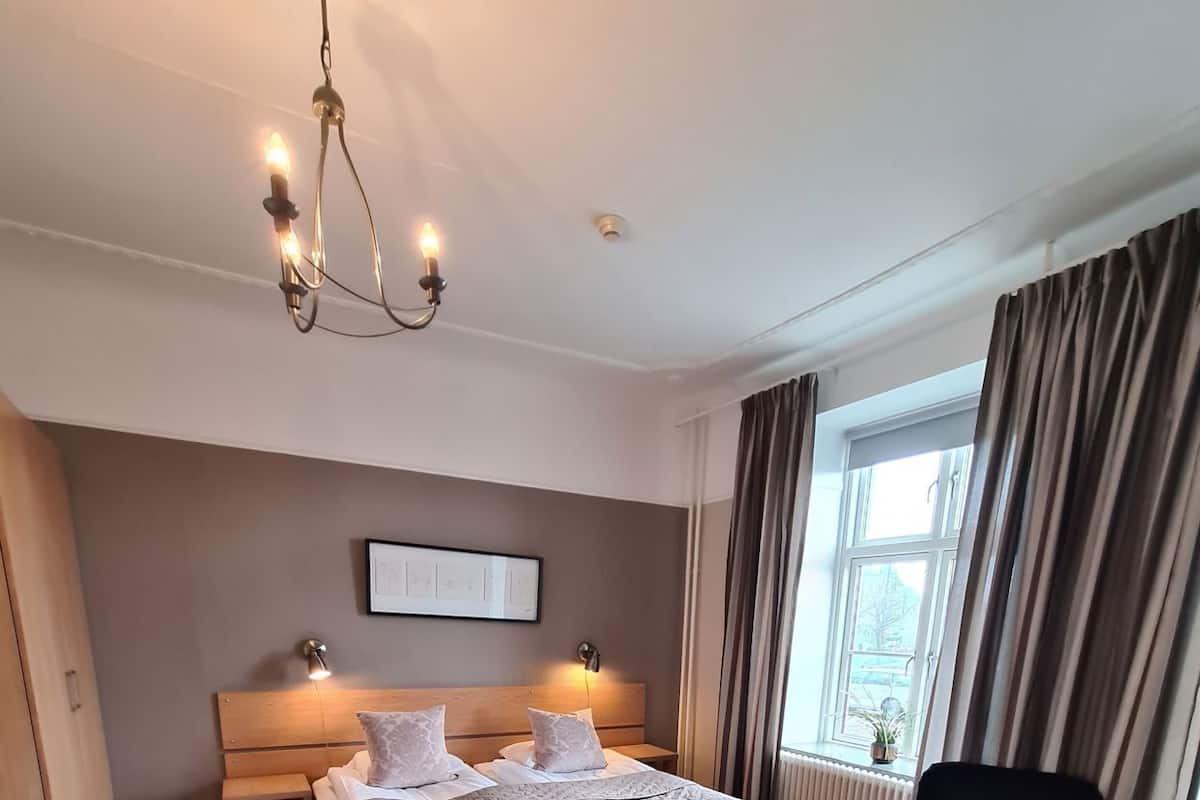 A bedroom with a bed, bedside tables, a chair, a window with curtains, a radiator, and a chandelier.