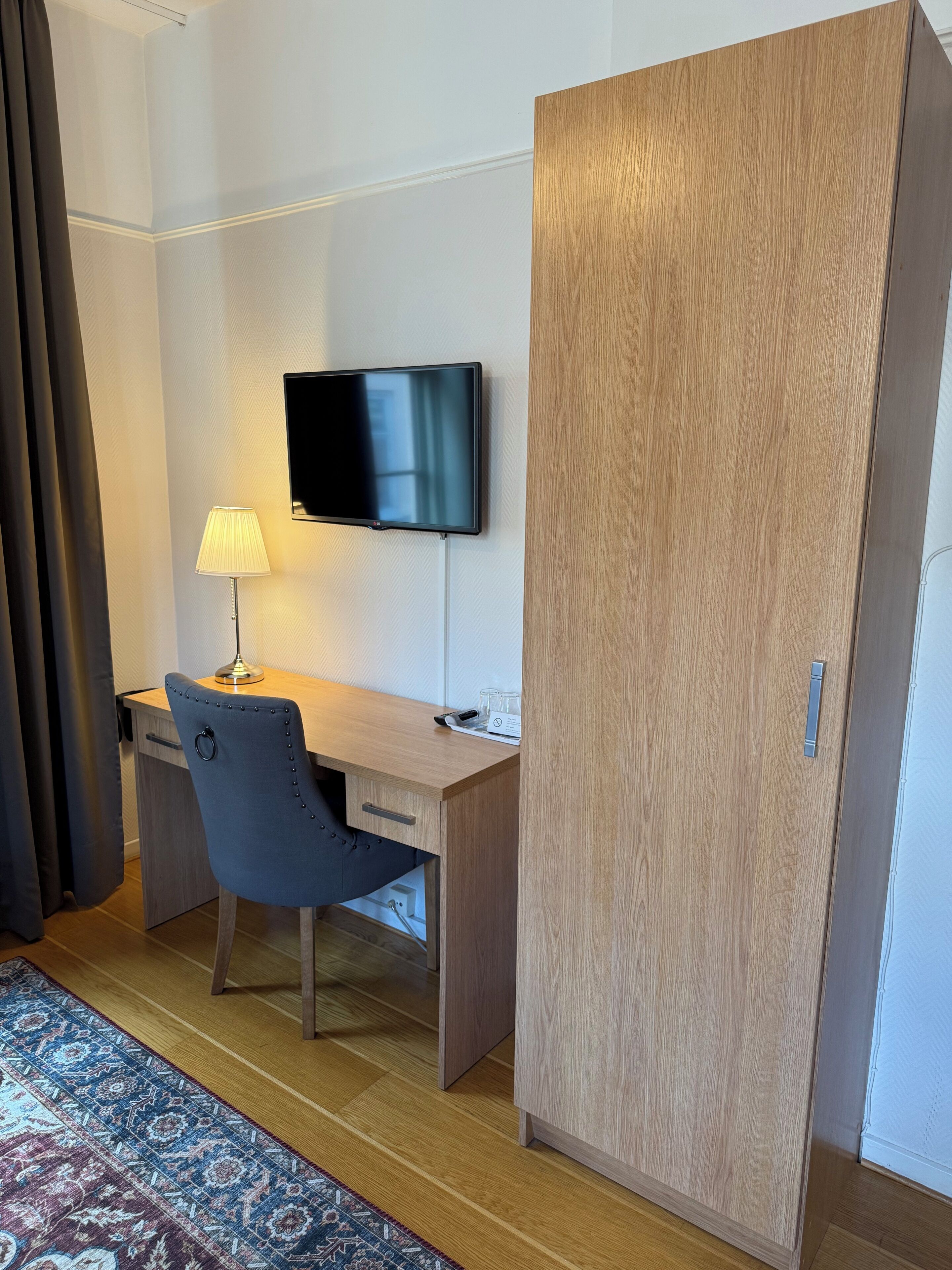 A hotel room with a wooden desk, a chair, a TV mounted on the wall, a lamp, and a wardrobe.