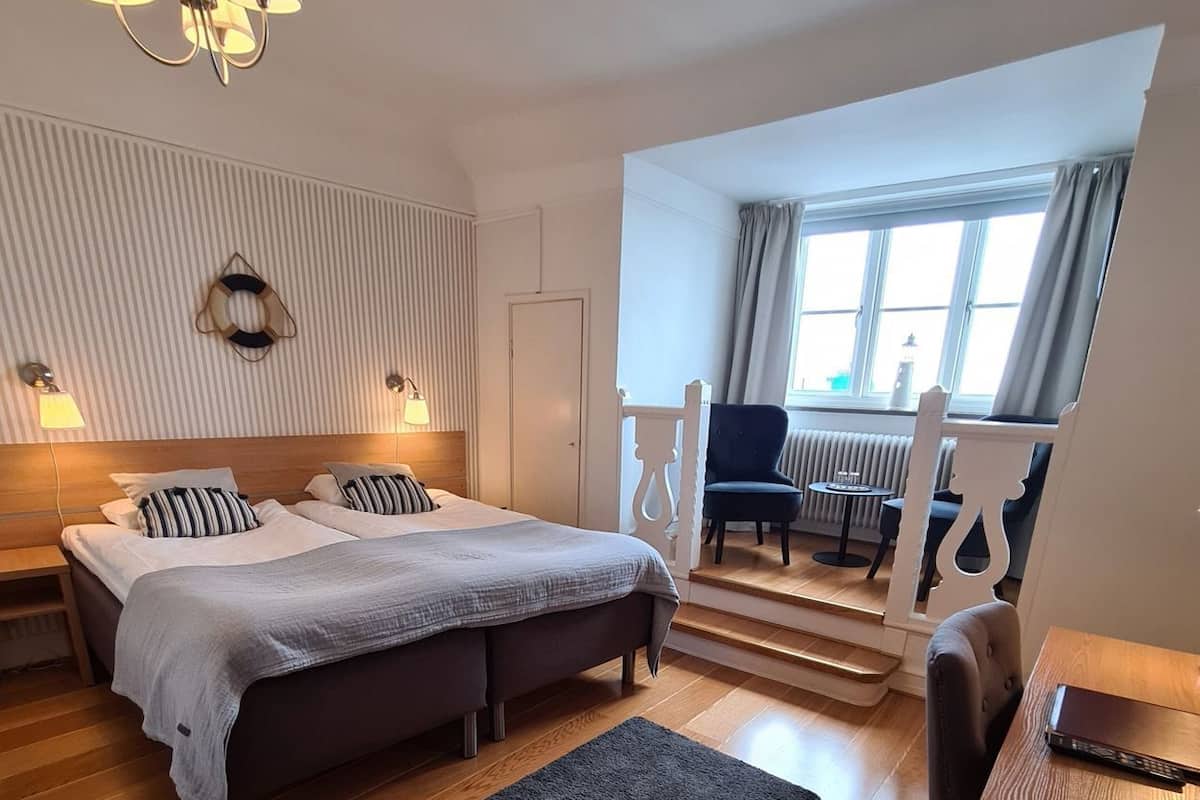 A hotel room with a bed, a desk, a chair, a small table, a lamp, a radiator, and a window with curtains.