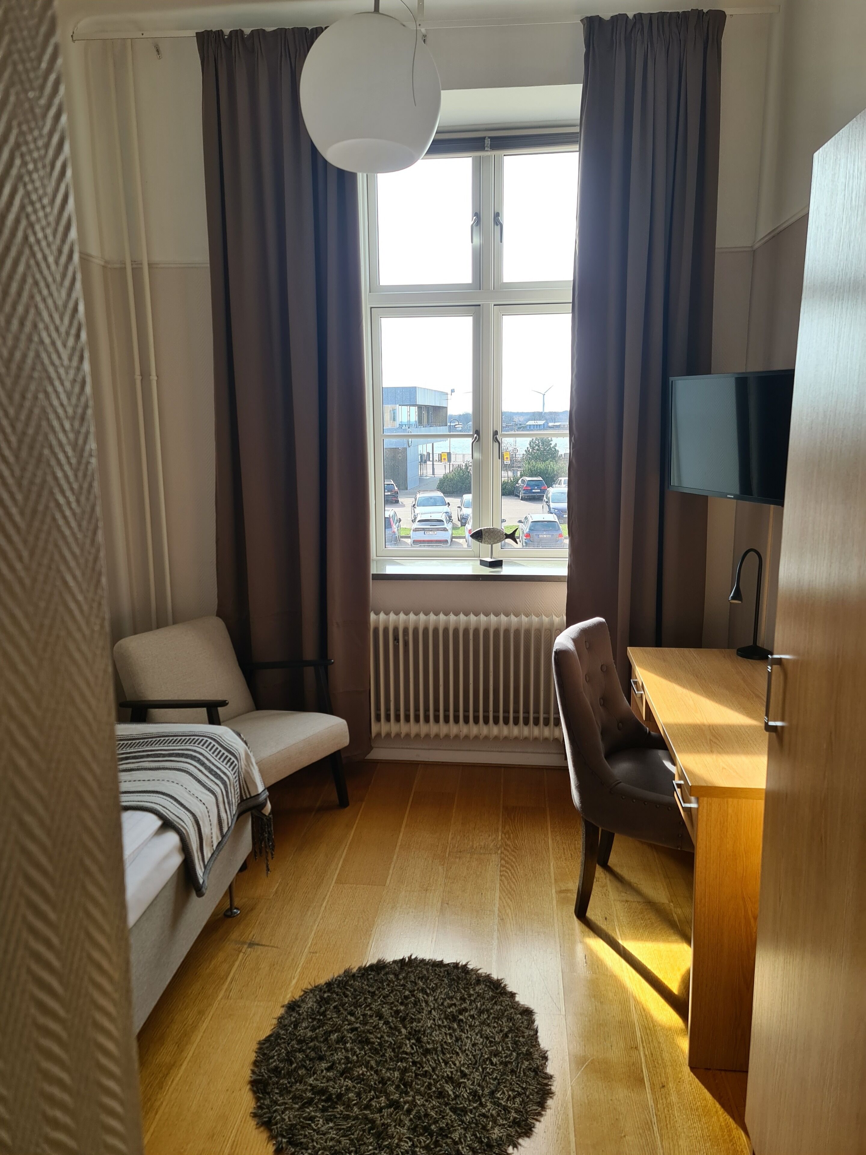 Ett litet, välstädat rum med en säng, ett skrivbord och en stol. Ett stort fönster ger utsikt över omgivningen, och ett element syns under fönstret.