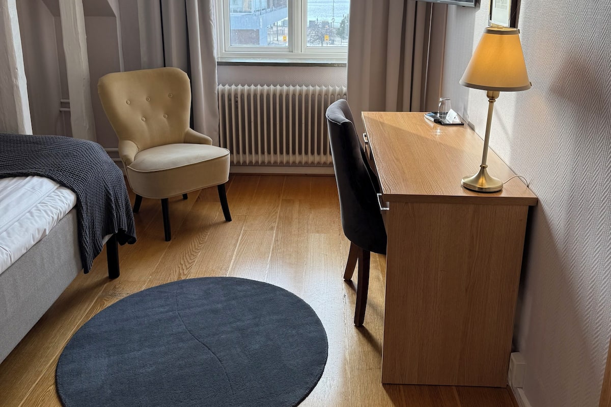 A hotel room with a bed, a desk with a lamp, a chair, a round rug, and a TV on the wall.
