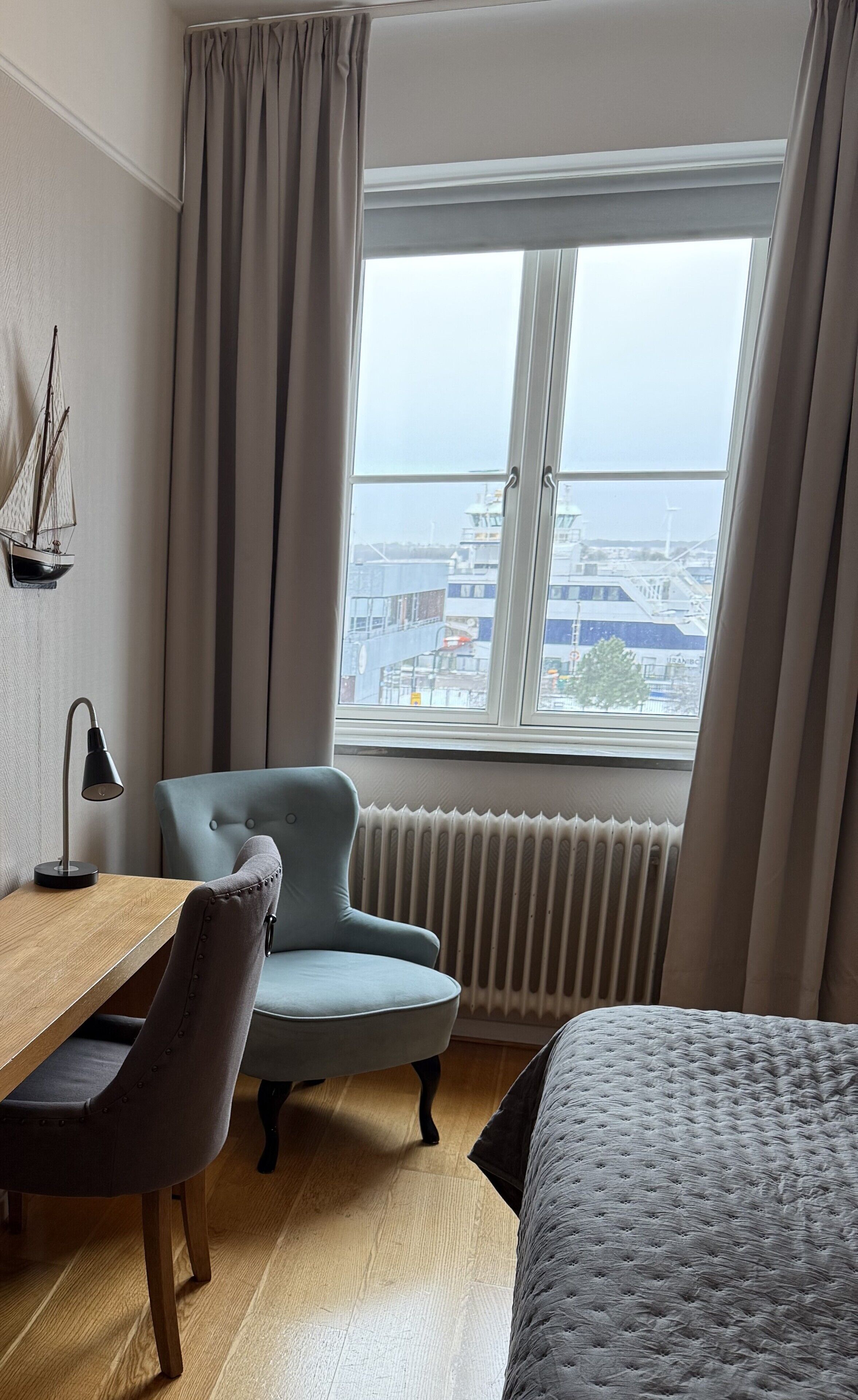 Una camera d'albergo con una scrivania in legno, una poltrona blu, un letto e un'ampia finestra con vista sulla città.