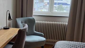 Una camera d'albergo con una scrivania in legno, una poltrona blu, un letto e un'ampia finestra con vista sulla città.